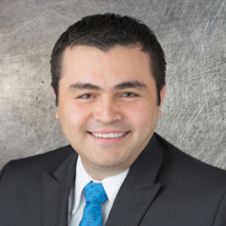 Professional headshot: smiling man in a dark suit and blue tie against a textured gray backdrop.