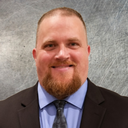 Portrait of a smiling man in a dark suit, light blue shirt, and patterned tie, against a gray textured background.