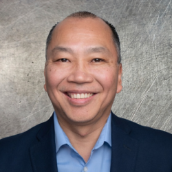 Portrait of a smiling man in a navy blazer and light blue shirt against a metal backdrop.