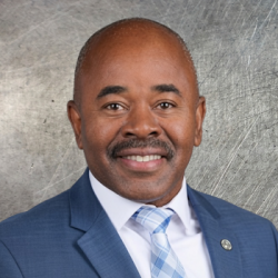 Head-and-shoulders portrait of a smiling man in a blue suit and light blue patterned tie against a gray backdrop.